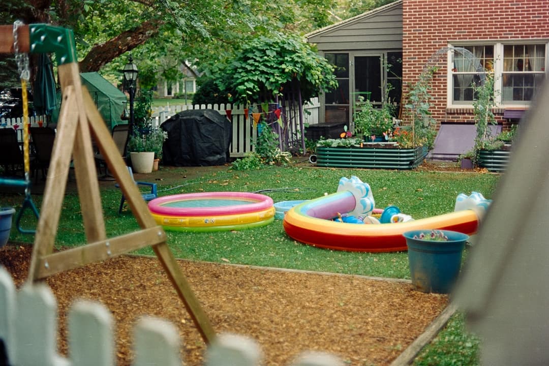 Backyard scene with colorful inflatable pools, swing set, and green lawn, summer fun