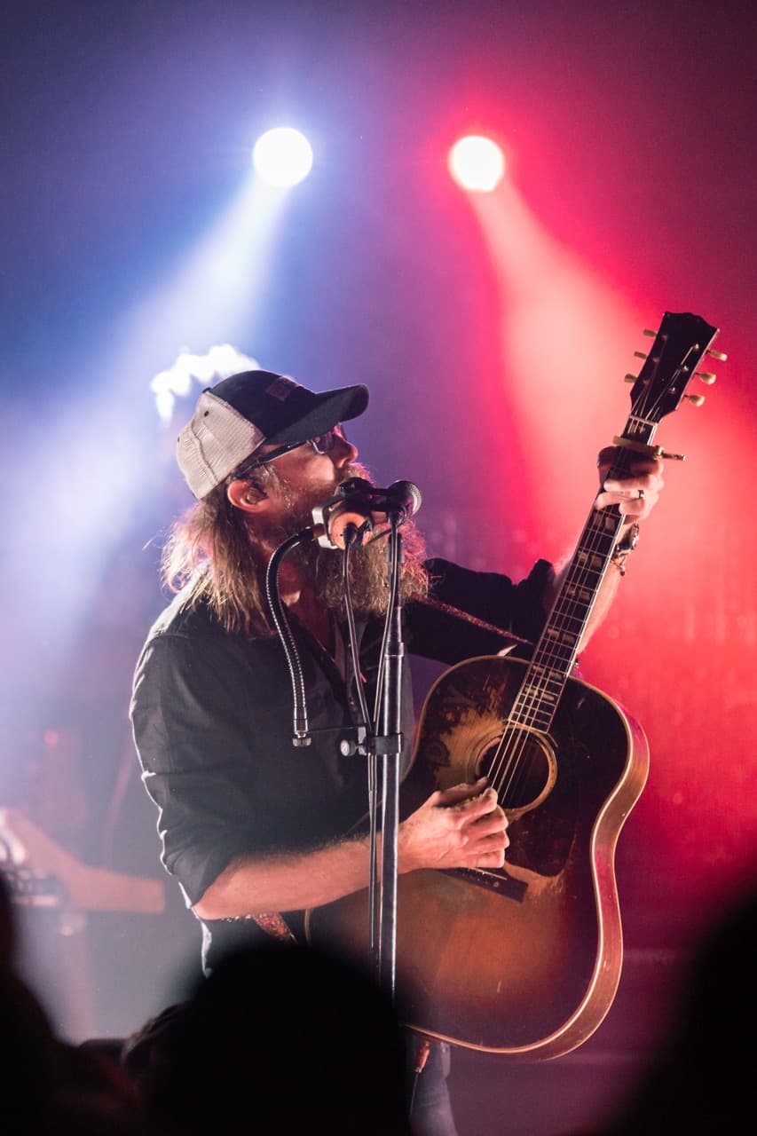 Live folk music performance: Singer-songwriter with beard playing acoustic guitar under red stage lights