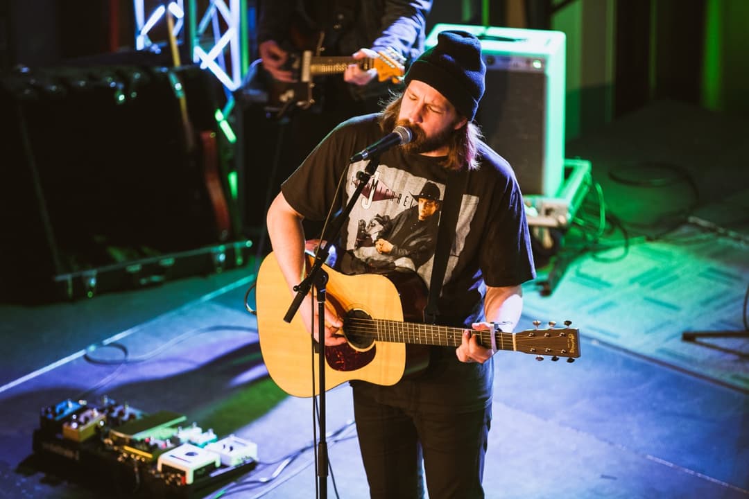 Live music performance: Musician with beard playing acoustic guitar on stage with green and blue lighting