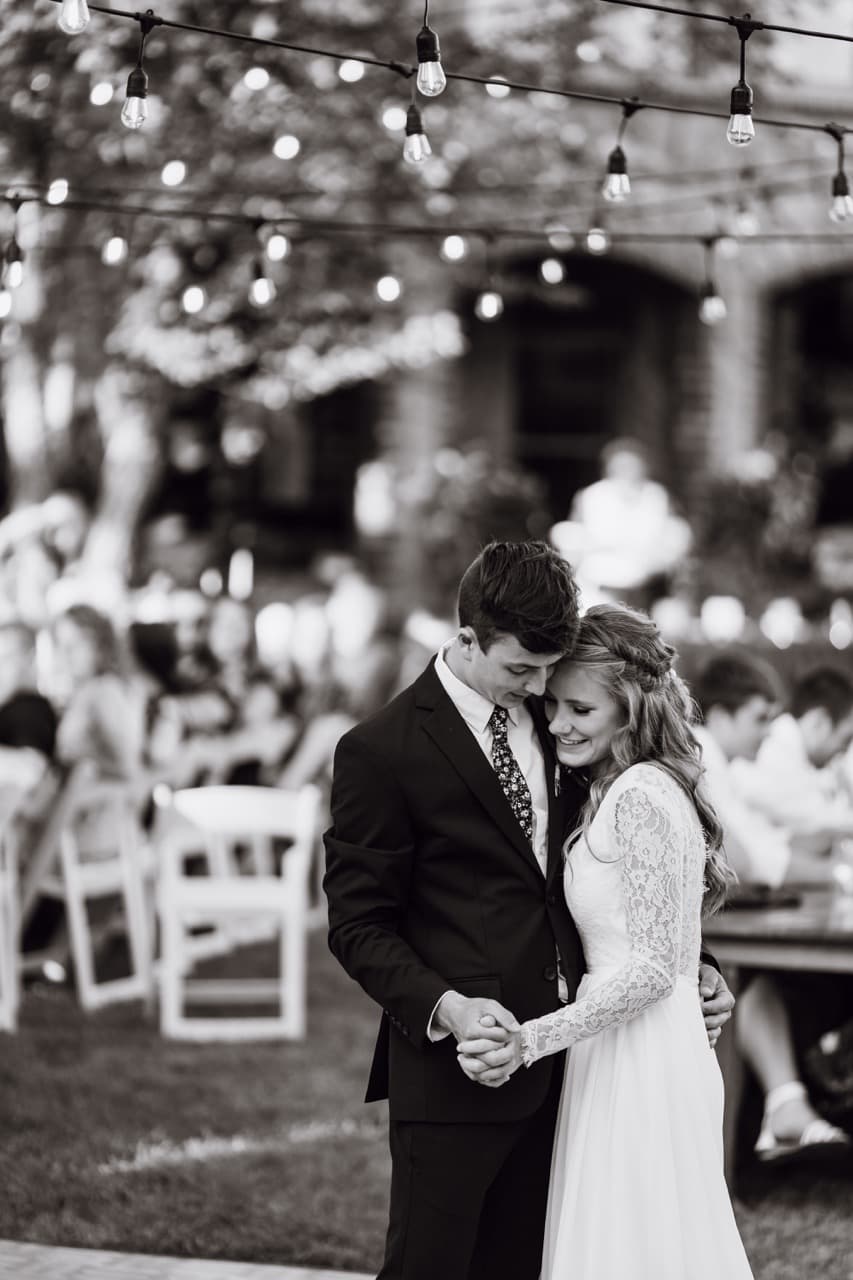 Black and white photo of a bride and groom dancing under string lights, romantic wedding moment