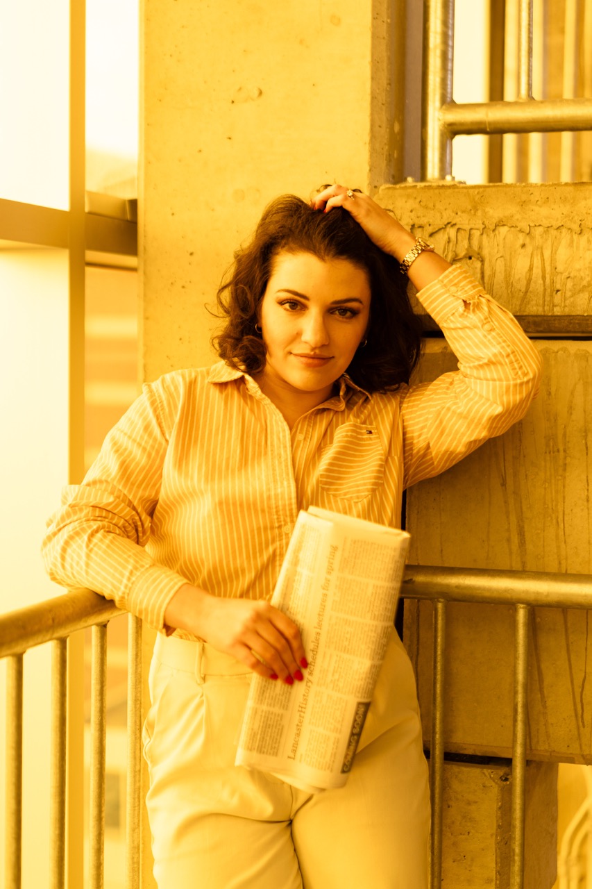 Model in cream-colored outfit leaning against sunlit wall holding newspaper